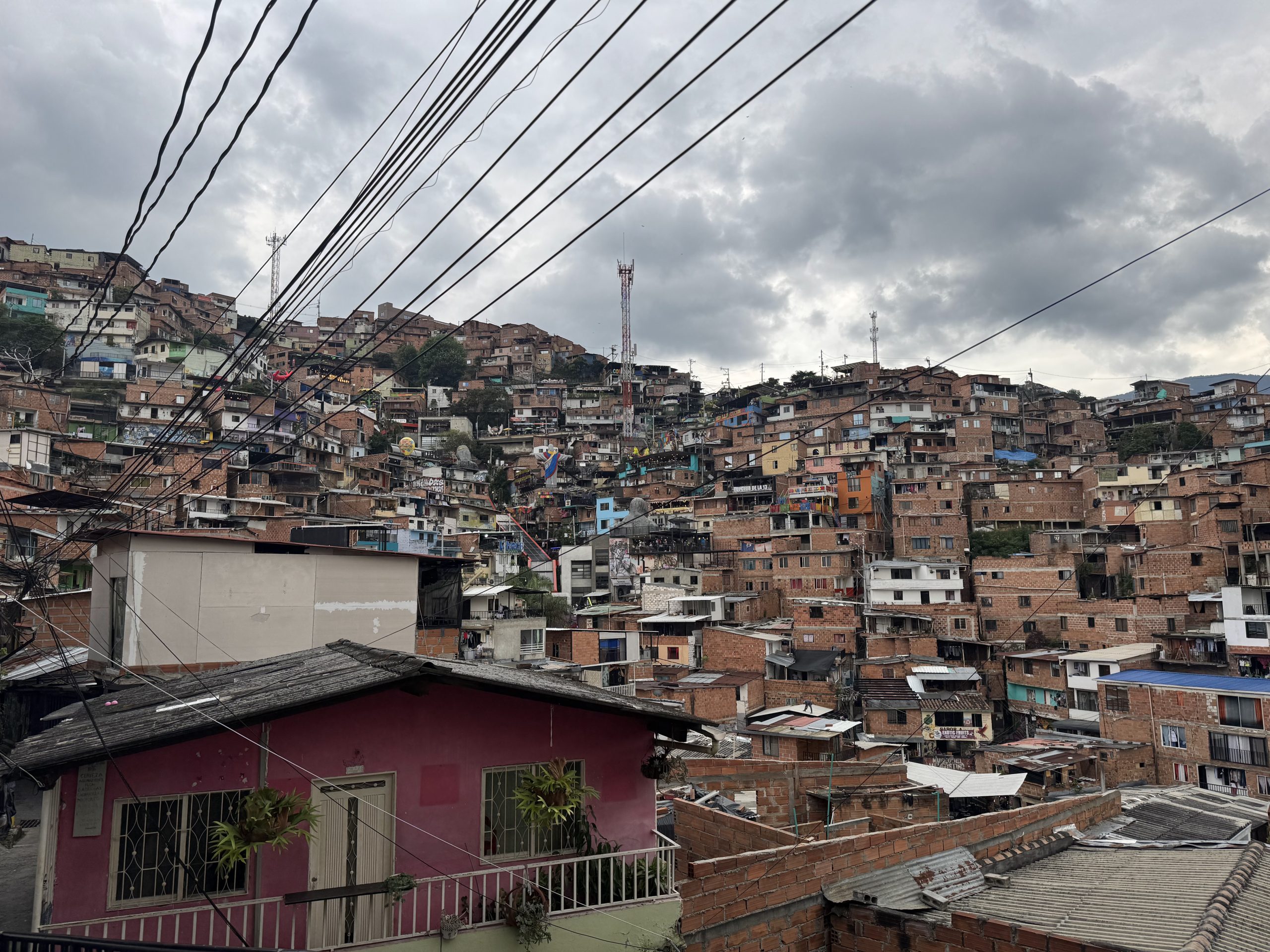 Photo of a densely packed hillside neighborhood with numerous small, closely built brick and concrete houses under a cloudy sky. Overhead power lines stretch across the scene