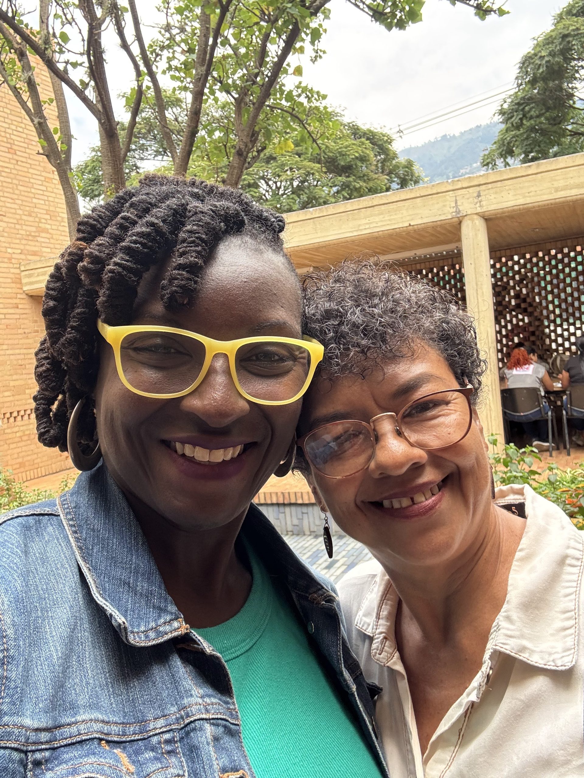 Photo of two smiling women posing closely outdoors with greenery and a brick building in the background. One woman wears bright yellow glasses and a denim jacket, while the other wears clear glasses and a light-colored shirt.