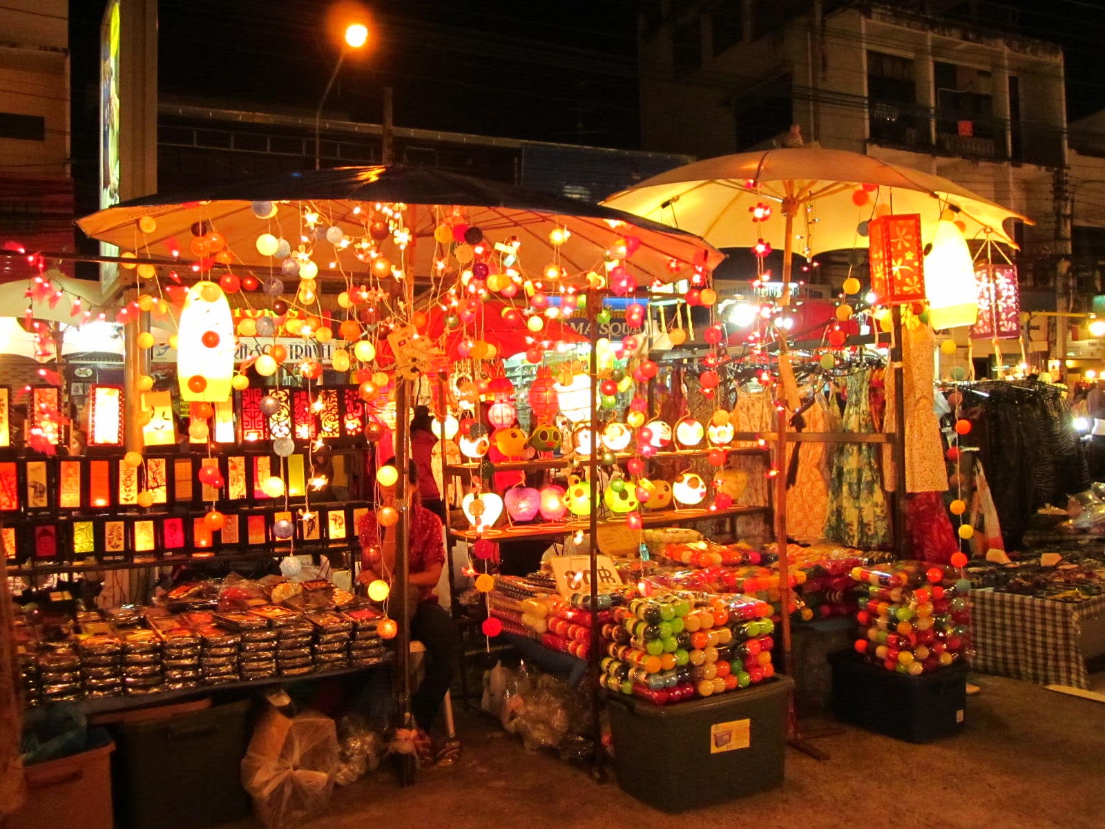 Photo of a vibrant night market stall decorated with numerous colorful hanging lanterns and lights, creating a warm, festive atmosphere. Various packaged goods and small items are displayed on tables beneath illuminated umbrellas, with surrounding buildings and streetlights visible in the background.