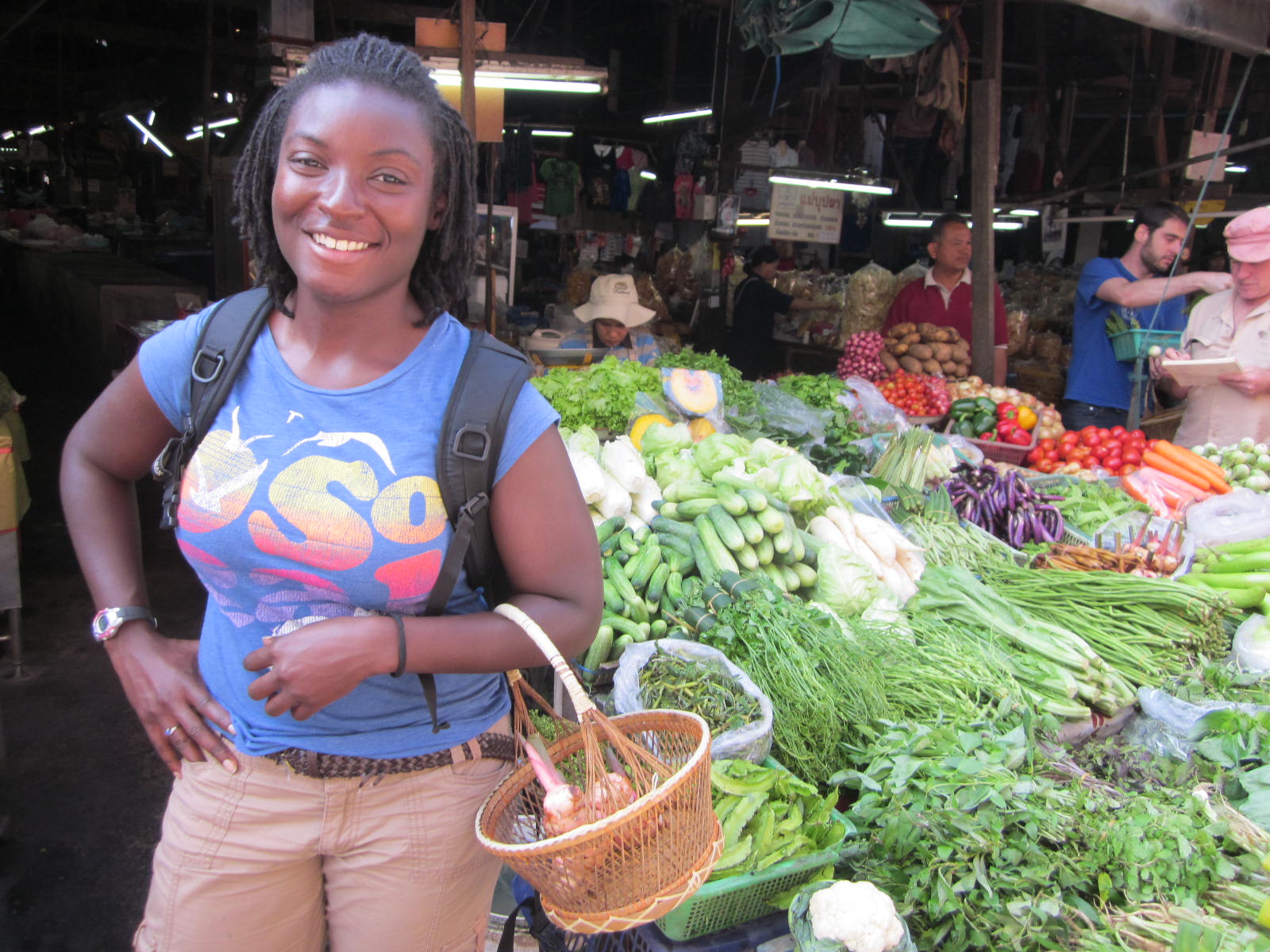 Photo of a smiling woman standing in front of a market stall filled with fresh vegetables, including cucumbers, eggplants, carrots, and leafy greens. The market setting is busy with other shoppers and vendors visible in the background