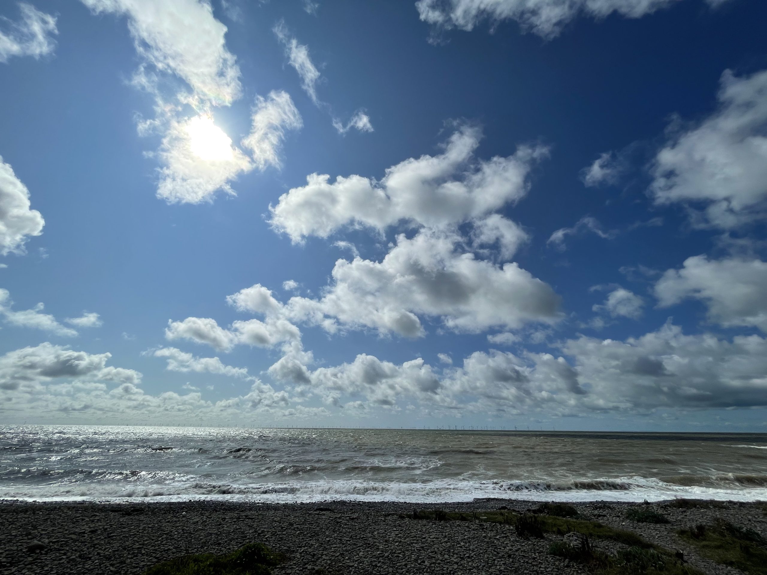Photo of a rocky shoreline and choppy ocean beneath a bright blue sky filled with scattered white clouds. Sun shines through clouds near upper left, while waves break across horizon and dark beach rocks occupy foreground.