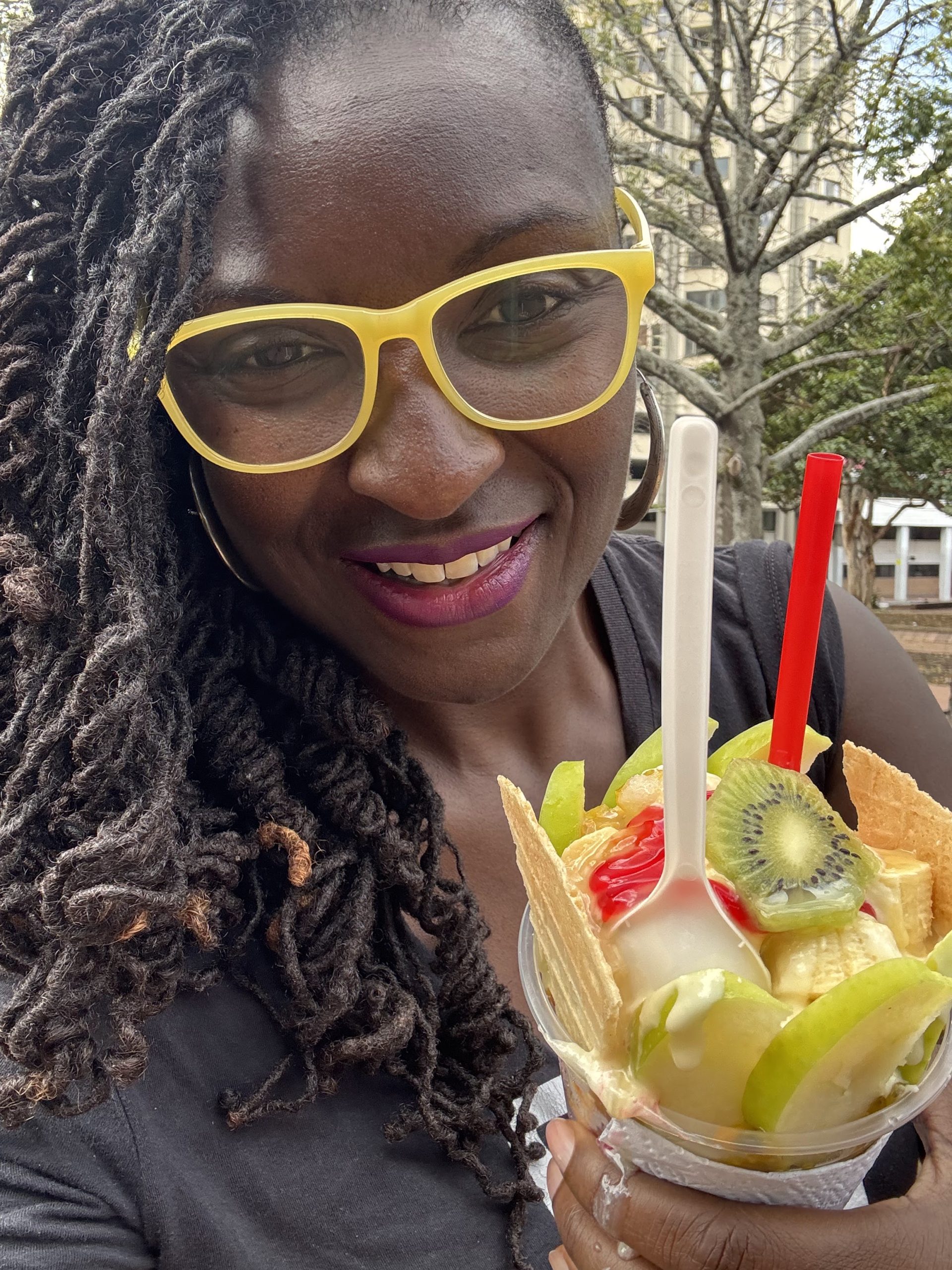 Photo of a smiling woman with long dreadlocks and yellow glasses holding a fruit-topped dessert in a waffle cone cup. Dessert includes kiwi slices, green apple slices, red syrup, and two spoons, set against an outdoor background with trees and buildings.