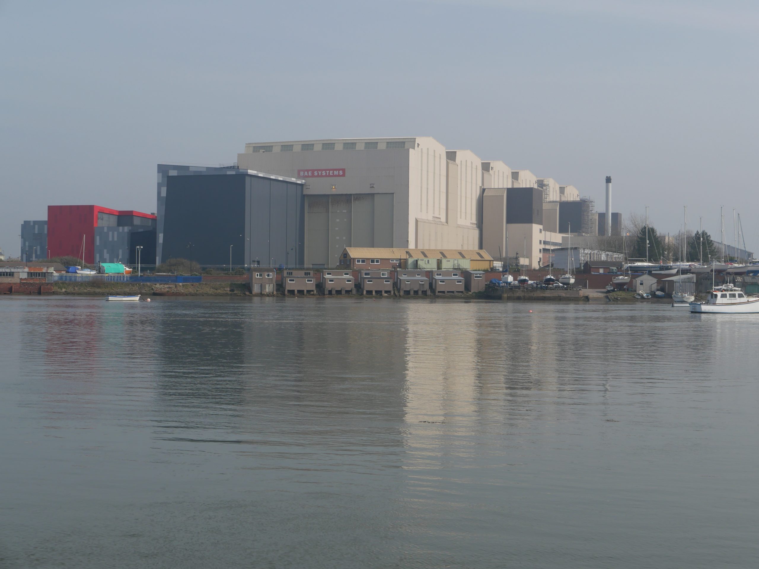 Photo showing a waterfront industrial complex with large gray and white factory buildings, smaller warehouses, and moored boats. Calm harbor water reflects the hazy buildings