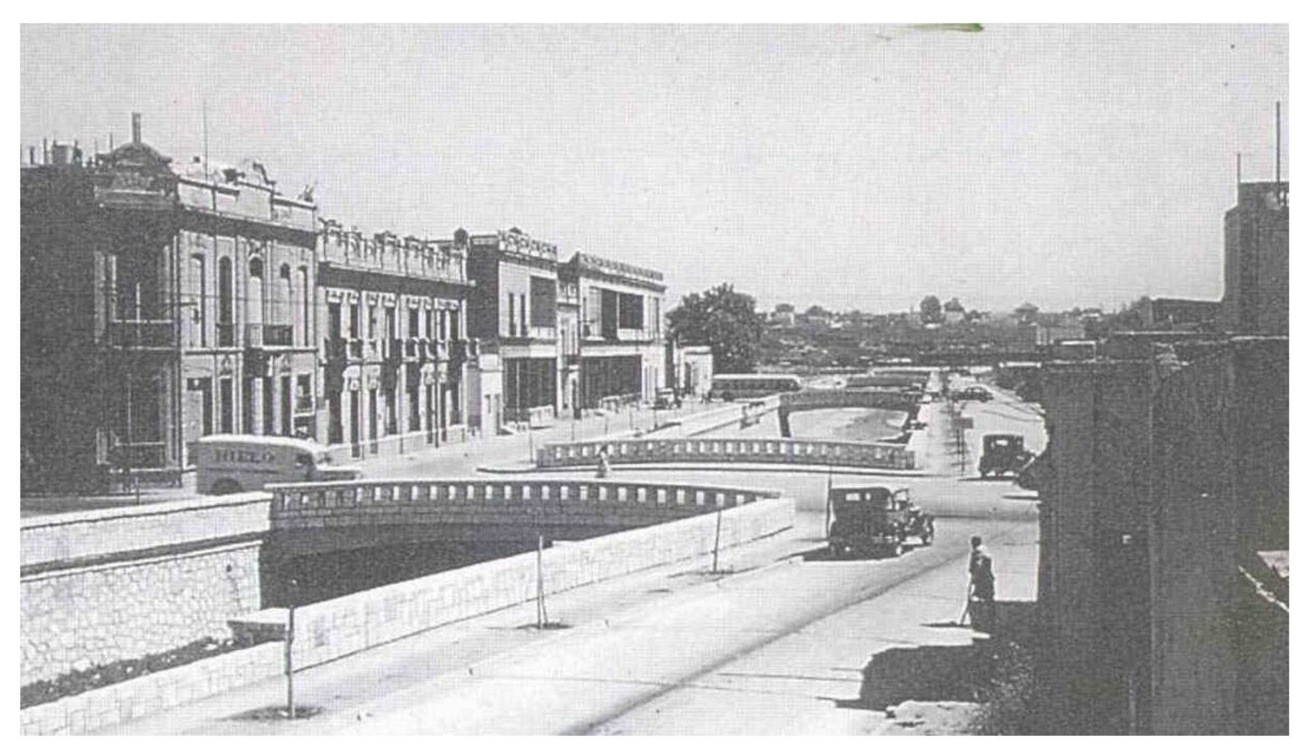 Historical black-and-white photo showing an urban roadway, bridge, and canal or river corridor lined with ornate early-20th-century buildings. Vintage cars, pedestrians, stone walls, streetlights, and distant low-rise structures fill the background