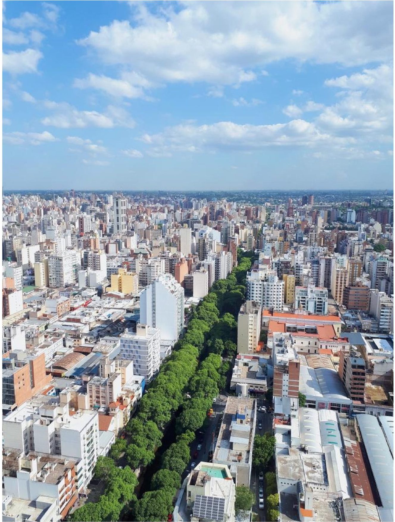 Aerial photo showing dense urban cityscape with high-rise buildings, low-rise structures, and a tree-lined boulevard extending toward horizon. Bright blue sky with scattered clouds frames the expansive metropolitan landscape