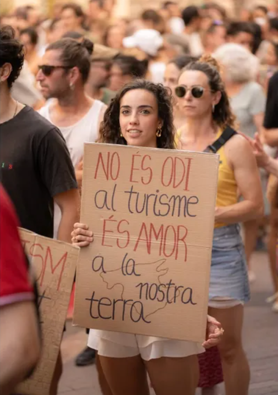 Photo showing a crowded street protest focused on tourism and local identity. Young woman holds a cardboard sign reading “No és odi al turisme, és amor a la nostra terra” (“It isn’t hatred of tourism; it’s love for our land”) amid demonstrators.