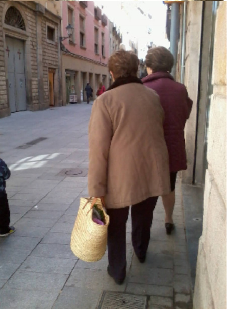 Photo of two older women walking along a narrow paved city street, one carrying a woven shopping basket. Historic stone and stucco buildings, pedestrians, sidewalk grates, and strong sunlight show an everyday urban scene.