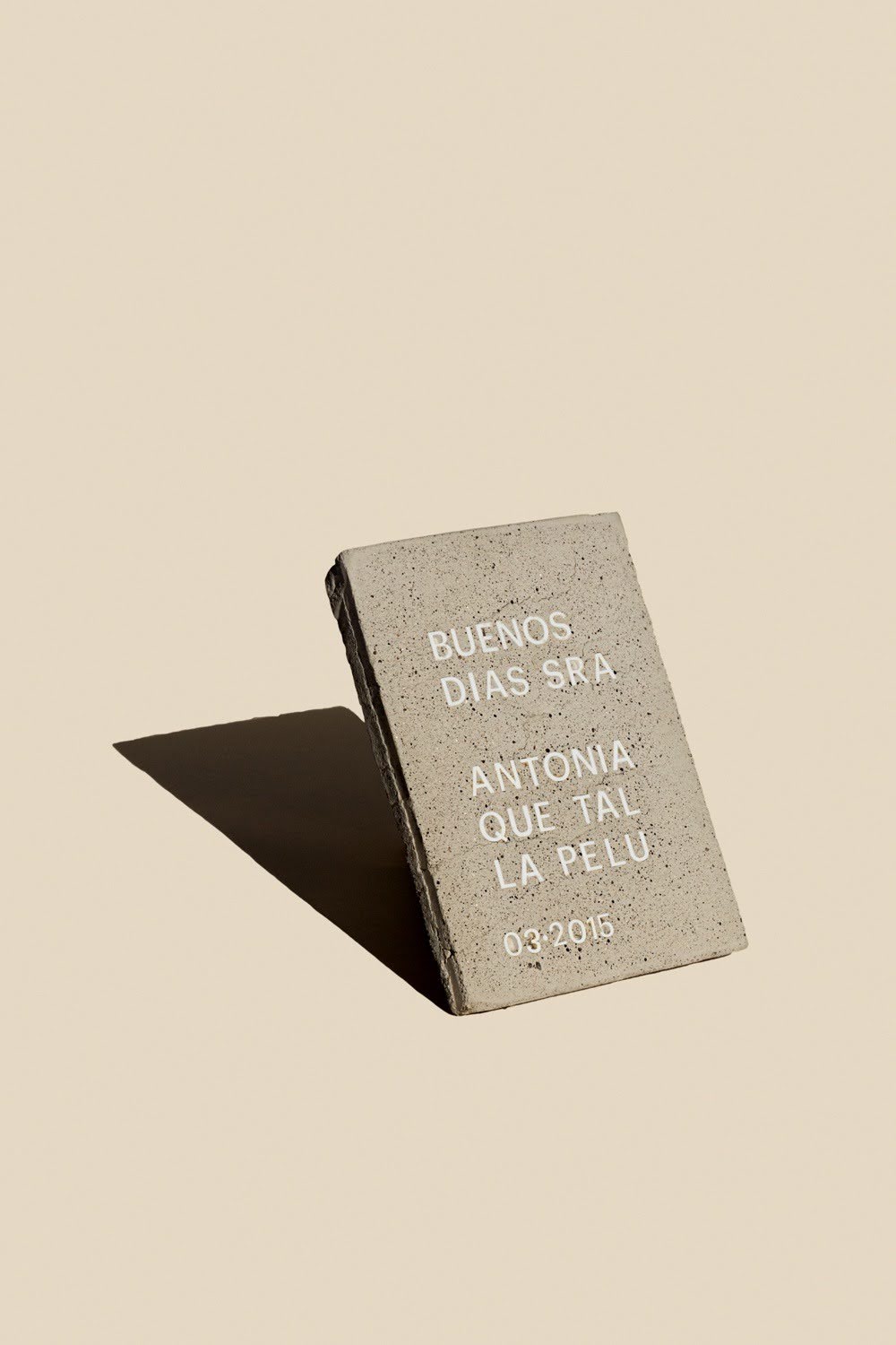 Photo shows a small gray, weathered book or plaque propped upright on a beige surface, casting a long shadow. White lettering reads “BUENOS DIAS SRA ANTONIA QUE TAL LA PELU” and “03-2015”. 