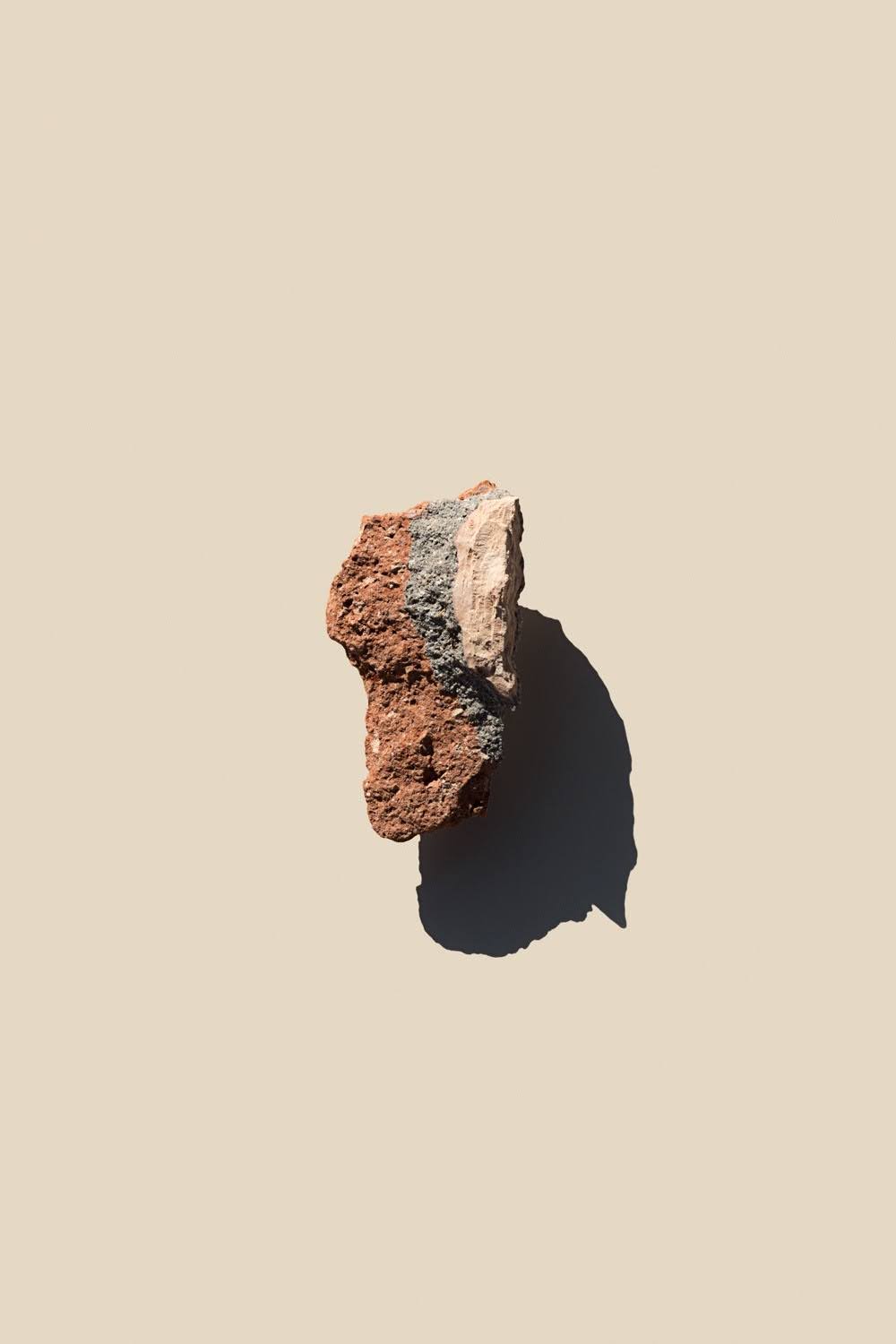 Photo documents an irregular reddish-brown and gray rock specimen isolated on a beige surface. Strong sunlight highlights rough, layered texture and casts a long dark shadow to lower right.