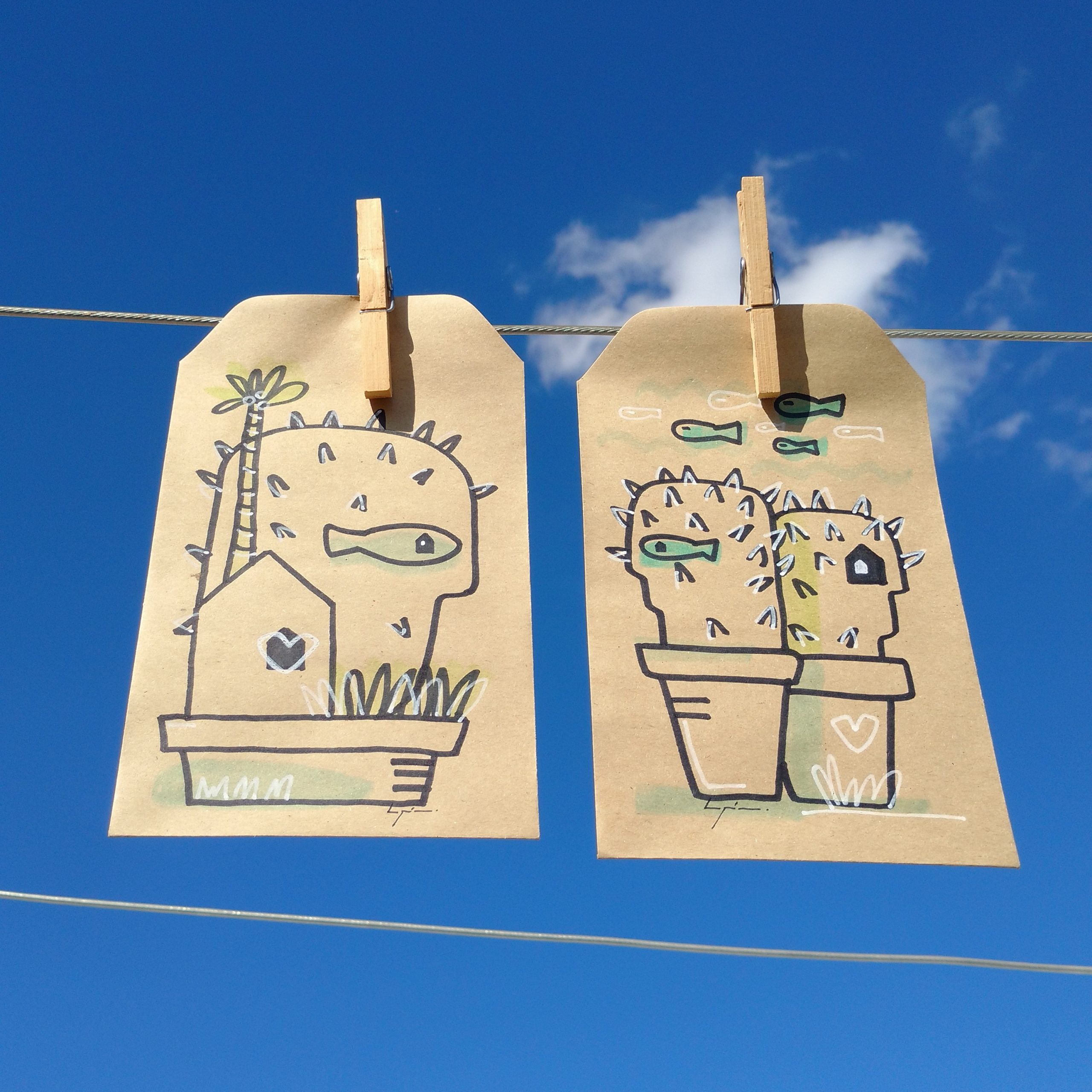Photo of two kraft-paper tags clipped to a clothesline against a bright blue sky. Left tag shows a potted, fish-holding creature beside a house, while right tag shows two spiky potted creatures with fish, hearts, and a small house.