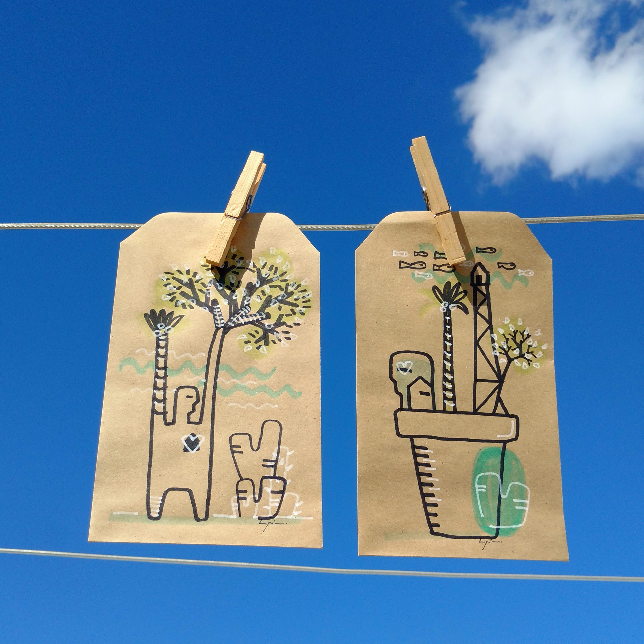 Photo of two kraft-paper tags clipped to overhead wires against a bright blue sky with clouds. Hand-drawn botanical scenes feature palm trees, cacti, planters, abstract markings, and decorative lines.