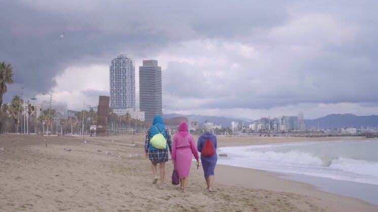 Photo showing three people walking along a sandy beach beneath dark storm clouds, with ocean waves, palm trees, and scattered birds. High-rise buildings, distant mountains, and a waterfront skyline form urban coastal backdrop