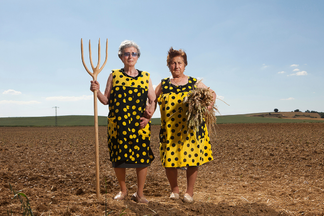 Photo showing two women standing in a freshly tilled field under a clear blue sky. One holds a pitchfork, while both wear matching black-and-yellow polka-dot dresses
