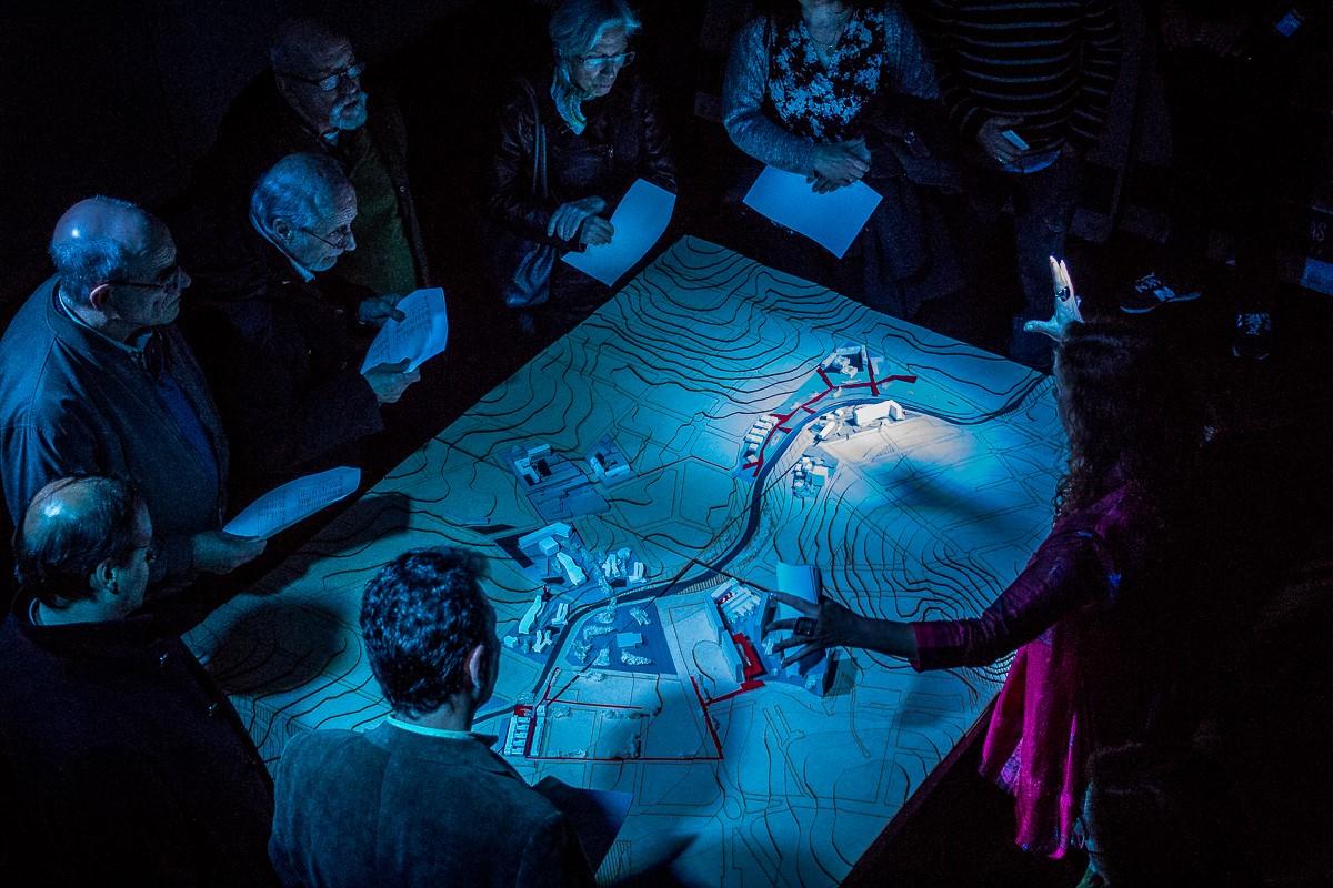 Photo shows a group gathered around an illuminated three-dimensional topographic map, apparently for geographic planning or educational discussion. Blue contour lines, miniature buildings, roads, and colored markers depict terrain and key locations, while participants hold notes and examine map features.