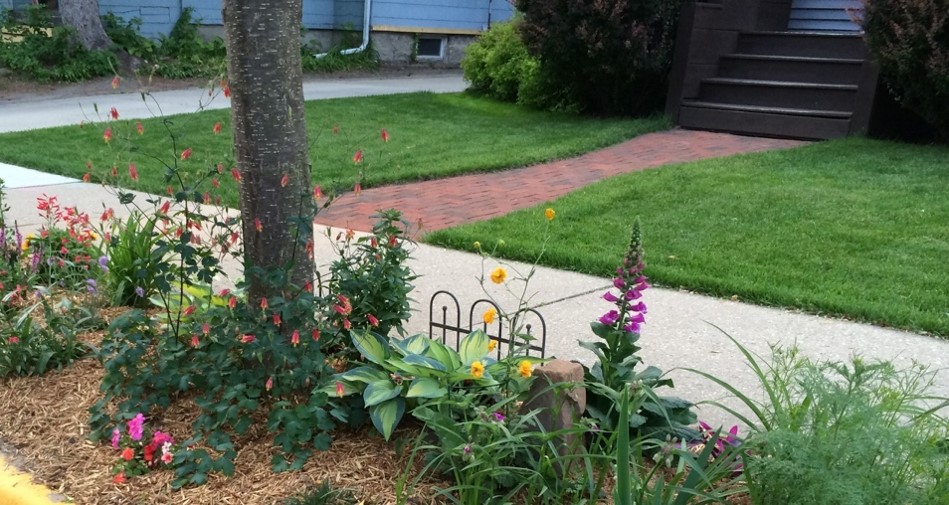 Photo showing a landscaped front yard with flower beds, mature tree, green lawn, and curved concrete walkway. Colorful red, pink, yellow, purple, and white flowers surround a small black metal garden border, with brick driveway and garage in background. 