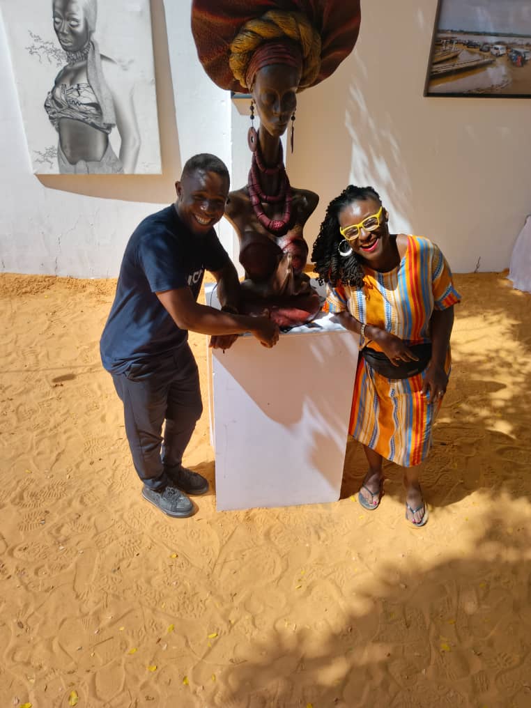 Photo of two people smiling and posing beside a detailed sculpture of a woman adorned with a large headwrap and jewelry, displayed on a white pedestal. The setting includes sandy ground, wall-mounted artwork, and sunlight casting shadows