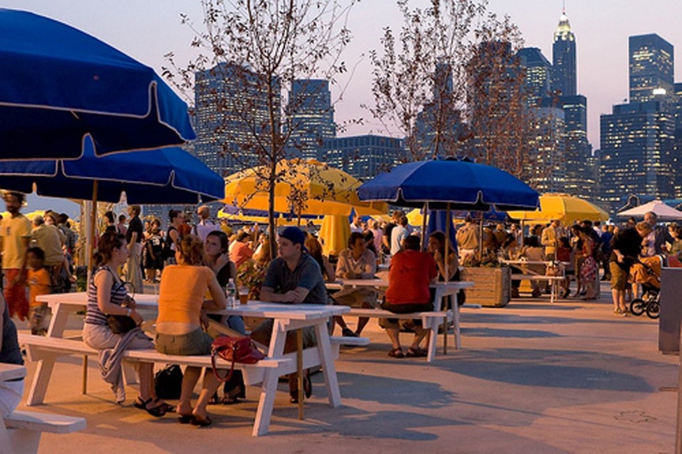 Photo of a vibrant, busy outdoor seating area with numerous people under blue umbrellas, with a backdrop of a city skyline at dusk.