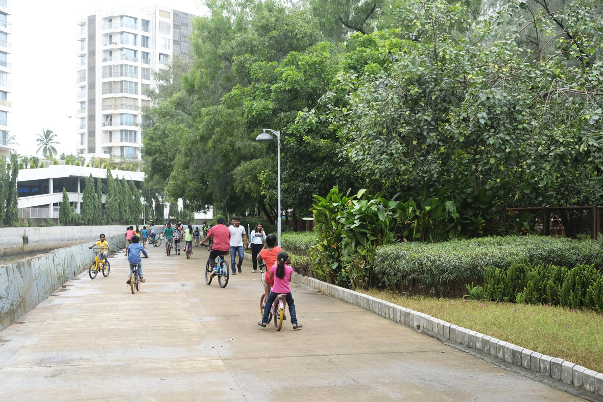 Photo of families and children cycling along a paved urban park path beside landscaped greenery and high-rise residential buildings. 