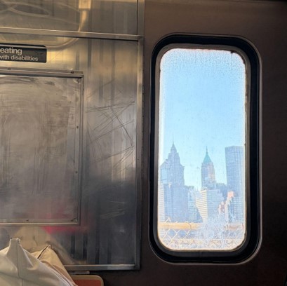 Photo of inside a subway car, showing a New York City skyline through a rounded window. Stainless-steel panel, partially visible “seating with disabilities” sign, worn seat, and blue-sky view featuring skyscrapers and bridge structure.