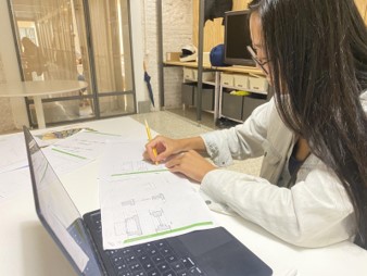 Photo of a woman marking architectural floor plans with a yellow pencil at a desk beside an open laptop. Office setting includes glass walls, shelving, monitor, scattered drawings, and green-highlighted plan details, suggesting design or drafting work.
