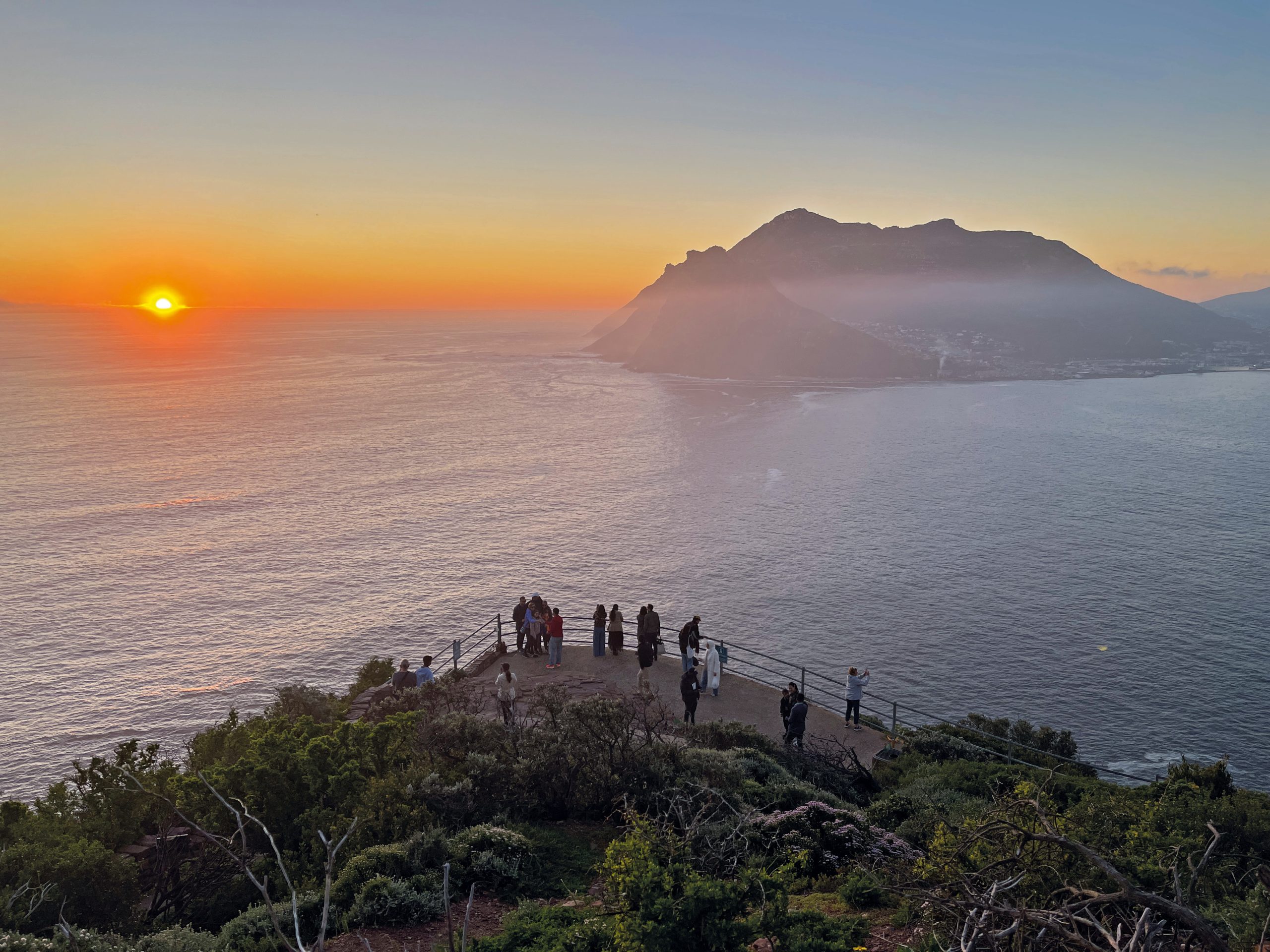 Photo of a sunset over a foggy ocean, with the bright orange sun near the horizon and mountain silhouette across bay. Viewpoint includes shrubs, railing, and visitors watching coastal scenery under clear blue-to-gold sky.