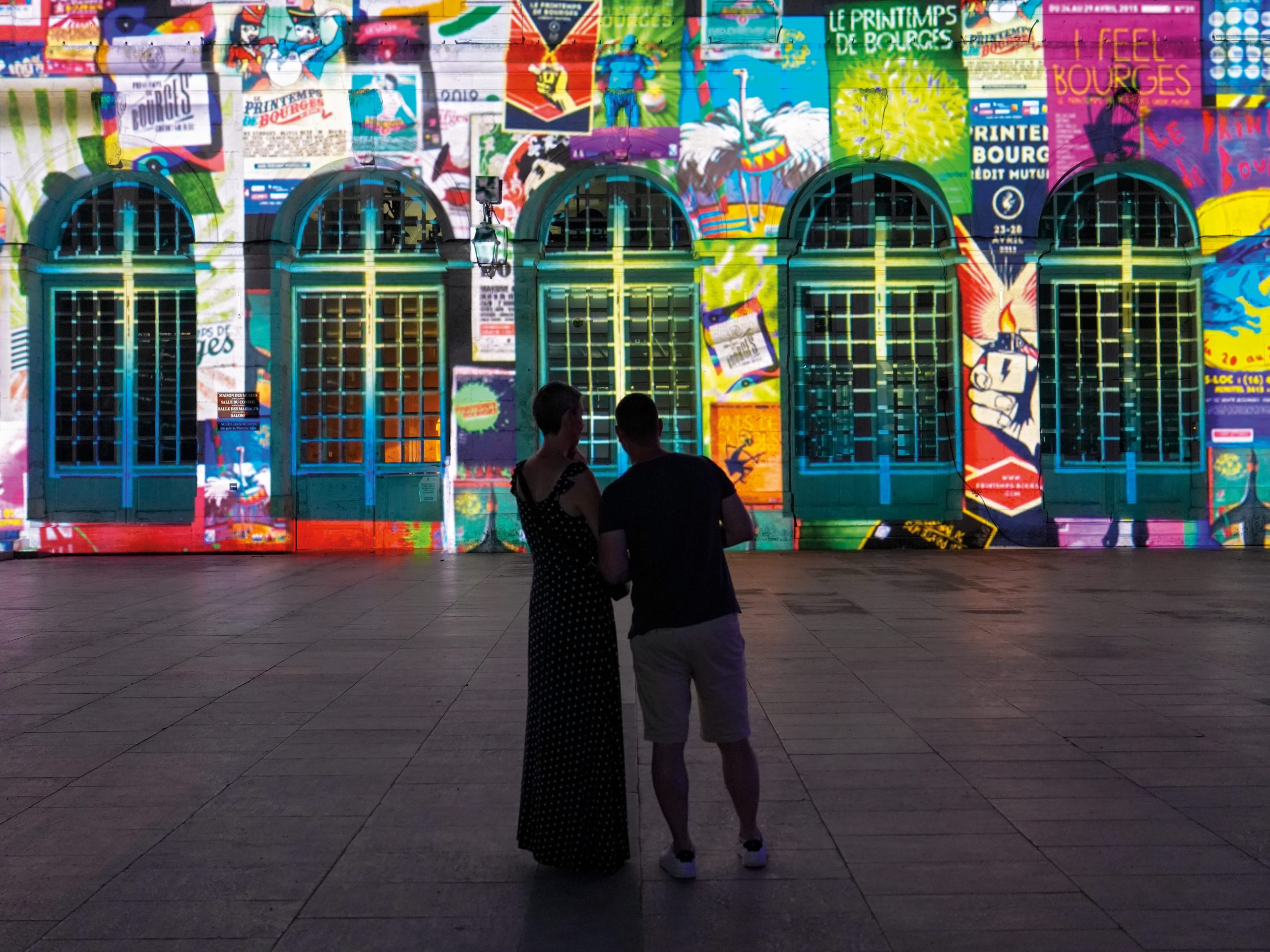 Photo showing a colorful, poster-covered building facade with five turquoise arched windows and doors. Two people stand in foreground, completely shadowed
