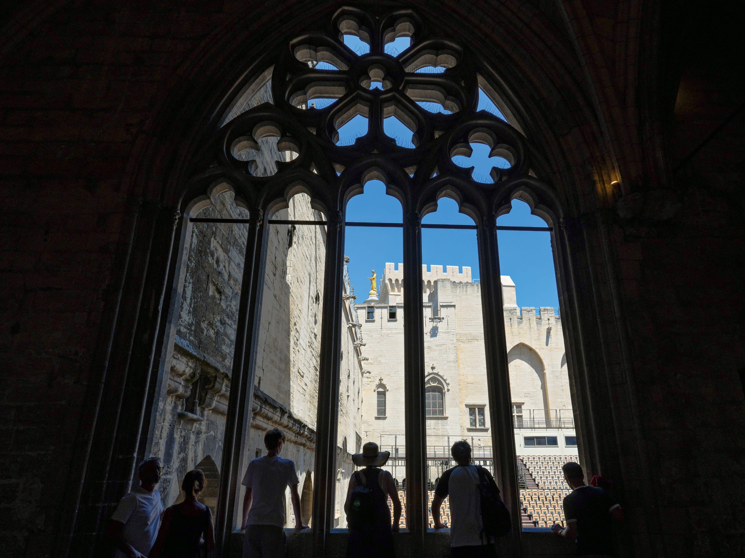Photo of a group of people silhouetted beneath an ornate Gothic open-air window, viewing a sunlit stone castle courtyard. Quatrefoil tracery, pointed lancet panels, crenellated walls, arched entry, and a bright blue sky fill the background